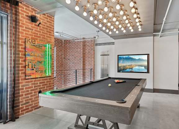 A pool table is in the middle of a room with a brick wall and large TV