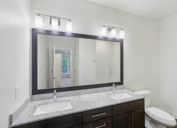 A bathroom with granite countertops, two sink, mirror, stand up glass panel shower, and toilet.
