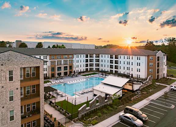 A sunset view of a hotel with a pool and cars parked in the parking lot.
