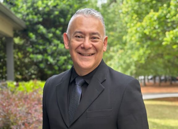 A man in a black suit and tie standing in front of a greenery.