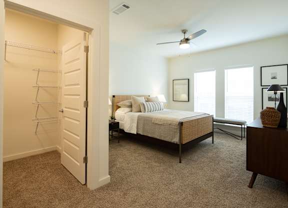 A bedroom with a bed, walk-in closet, dresser, windows, and a ceiling fan.