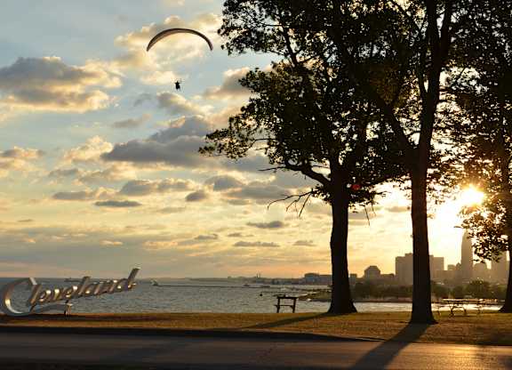 A person is paragliding over a park with trees and a Cleveland sign in the background.