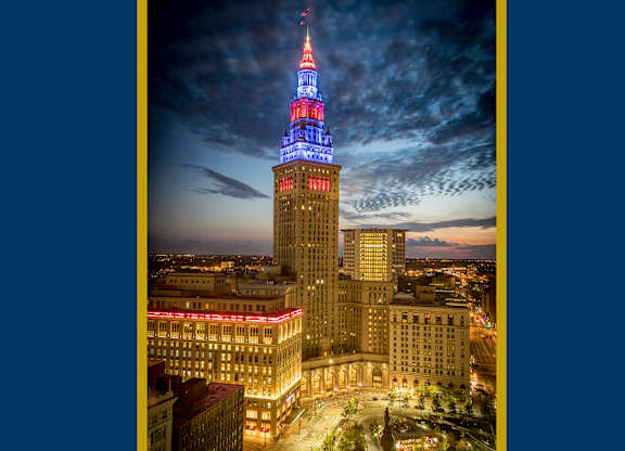 High-angle twilight view of The Terminal Tower Residences illuminated in patriotic red, white, and blue lights, soaring above the cityscape of downtown Cleveland.