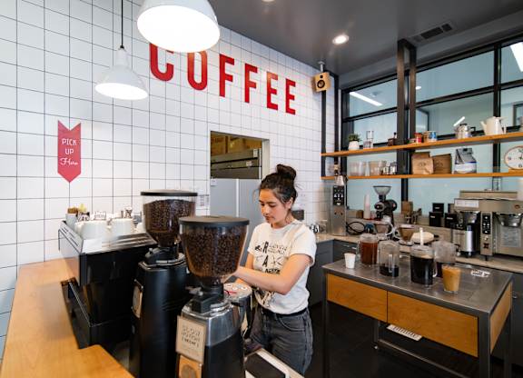 A woman making coffee with a white wall behind her that says coffee.