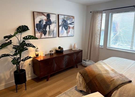 A bedroom with a bed, a plant, a wooden cabinet, and two paintings on the wall.