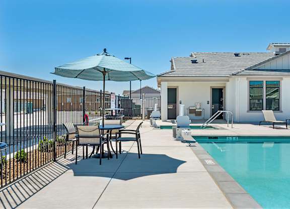A poolside table with chairs and an umbrella is in front of a house.
