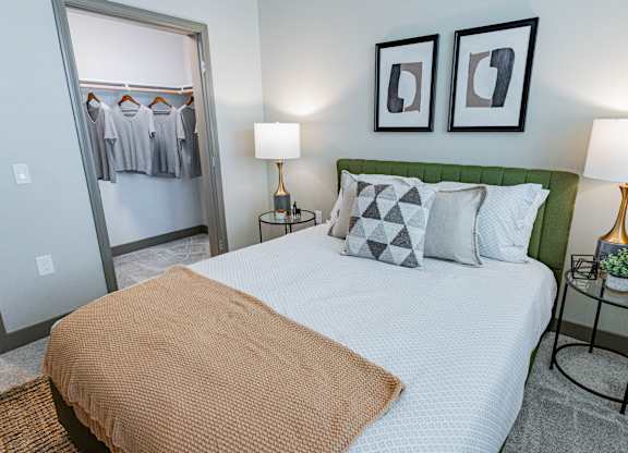 2-Bedroom Luxury Apartments In Burlingame, CA - Bayswater Burlingame - A Modern Bedroom With An Olive Green Tufted Headboard, White Patterned Bedding, And A View Into A Spacious Closet.