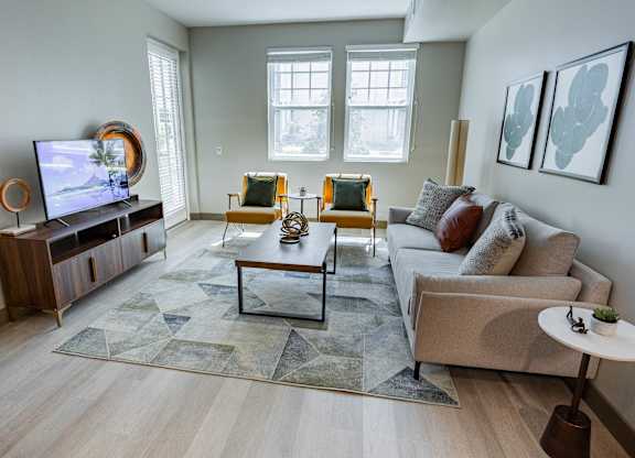 1-Bedroom Luxury Apartments In Burlingame, CA - Bayswater Burlingame - A Modern Living Room With A Grey Sofa, Yellow Accent Chairs, A Geometric Rug, And A Large Flat-Screen Television.