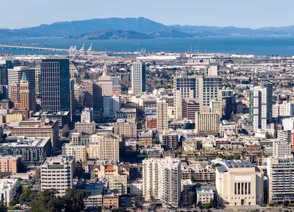 A cityscape with a mix of modern and older buildings, a bridge in the distance, and a mountain range in the far background.