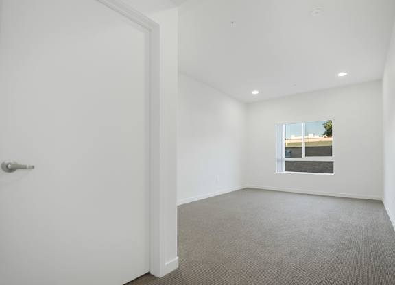 A white door is on the left of a room with a carpeted floor and a window with a view of a building.