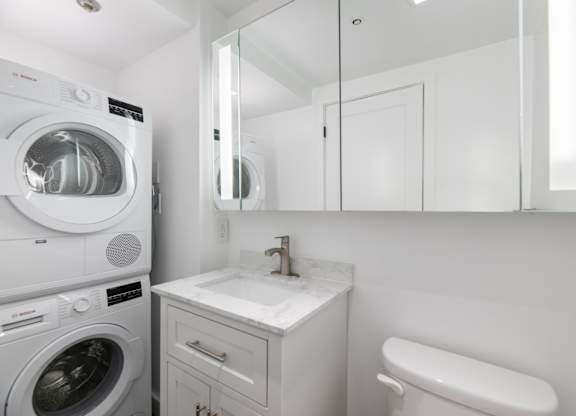 A white washing machine is in a small laundry room with a sink and mirror.