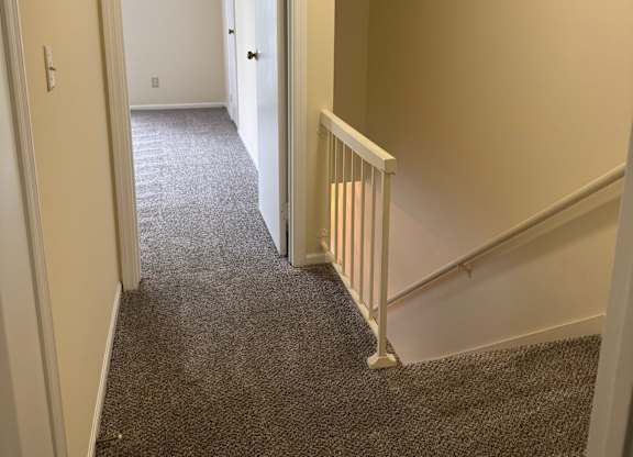 A hallway with a carpeted floor and a staircase with white railings.
