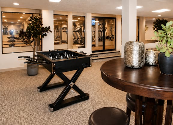 A foosball table is in the middle of a carpeted room with a round table and two potted plants.