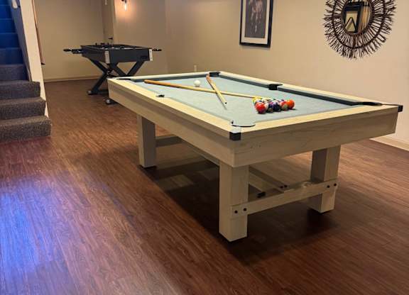 A pool table with a green cloth and white cues is in the middle of a room with a wooden floor and a mirror on the wall.