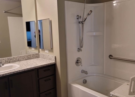 White tub/shower combo in bathroom, with dark wood cabinets. Tub has grab bars.
