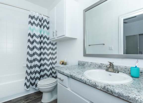 A bathroom with a white toilet, a grey and white shower curtain, a marble countertop, and a white sink.