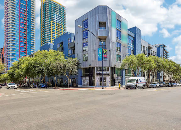 A modern building with a blue and grey facade is in the foreground of a street with cars parked on the side.