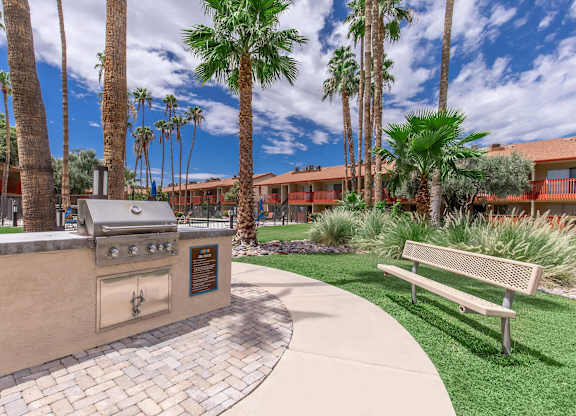 A park with a bench, a grill and palm trees.