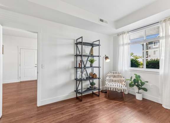 A room with a wooden floor, a chair, a shelf with plants and decorative items, and a window with white curtains.