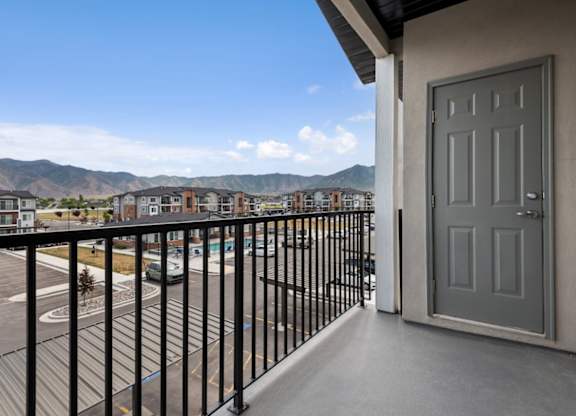 A balcony with a grey door and a black railing overlooking a parking lot and houses.
