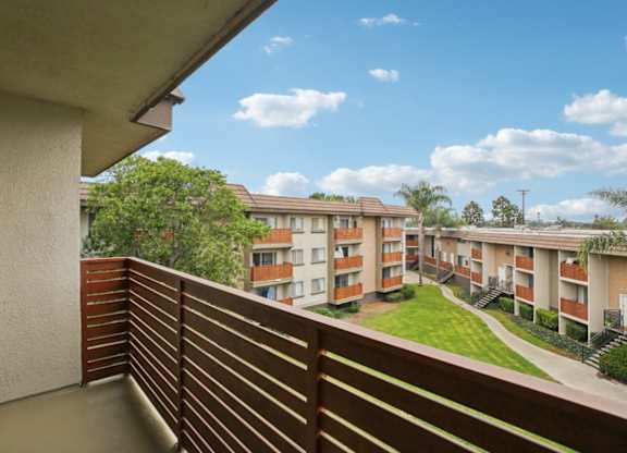 A balcony with a wooden railing overlooks a grassy courtyard and apartment buildings.