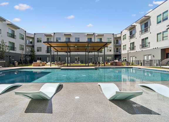 A swimming pool with sun loungers in front of apartment buildings.