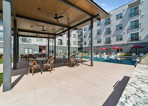 A patio with a table and chairs is surrounded by a pool and buildings.