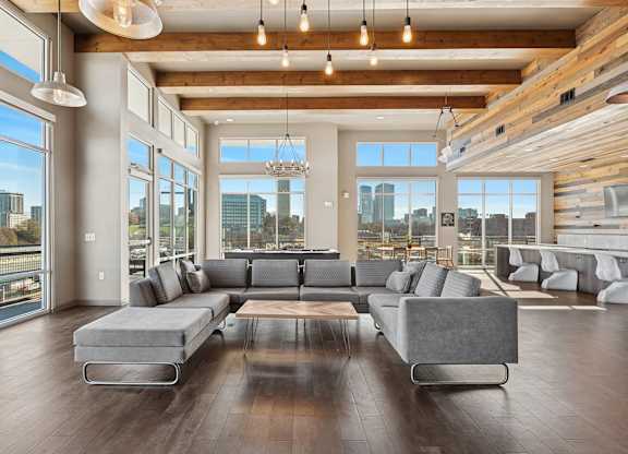 A large grey couch sits in the middle of a spacious room with wooden floors and a city view through the windows.