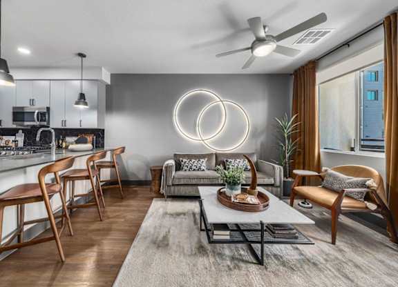 A modern living room with a grey rug, a grey sofa, a grey coffee table, a grey wall, a grey ceiling, a grey floor, a grey kitchen, a grey dining table, a grey chair, a grey plant,