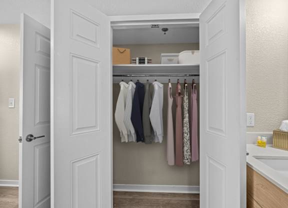 A white door opens to a small closet with clothes hanging on a rod.