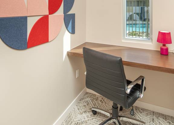 A red, blue and grey abstract art piece hangs on a wall above a desk with a black chair.