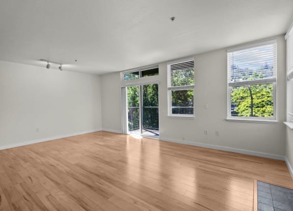 Spacious and bright living room with white walls, wood-style flooring, overhead lighting, and sliding glass door leading to private balcony at Collins Circle apartments in the Goose Hollow district in Portland, Oregon.