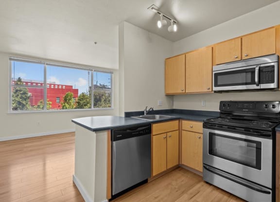 Kitchen with stainless steel appliances, wood cabinets, and wood-style flooring looking into living room area with window at Collins Circle apartments in the Goose Hollow district in Portland, Oregon.