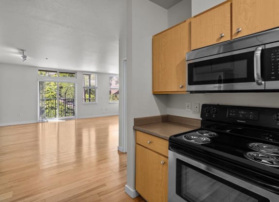 Kitchen with stainless steel appliances, wood cabinets, and wood-style flooring showing view into living room with large windows at Collins Circle apartments in the Goose Hollow district in Portland, Oregon.