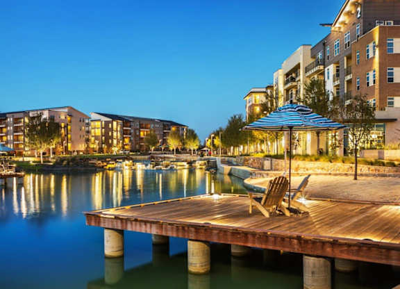 A wooden dock with chairs and an umbrella is in the foreground of a cityscape with buildings and boats.