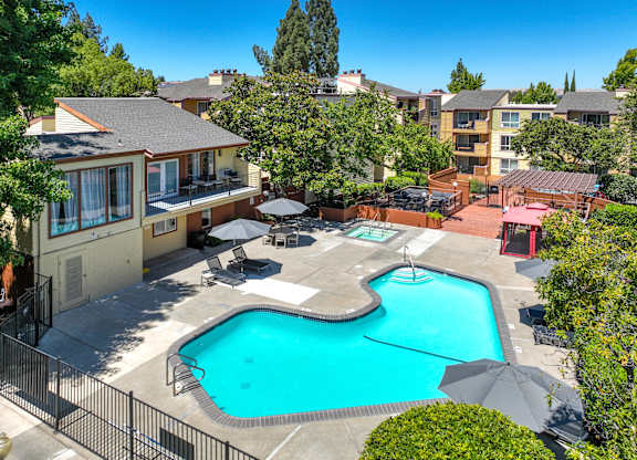 A swimming pool surrounded by a black fence and a patio with chairs and an umbrella.