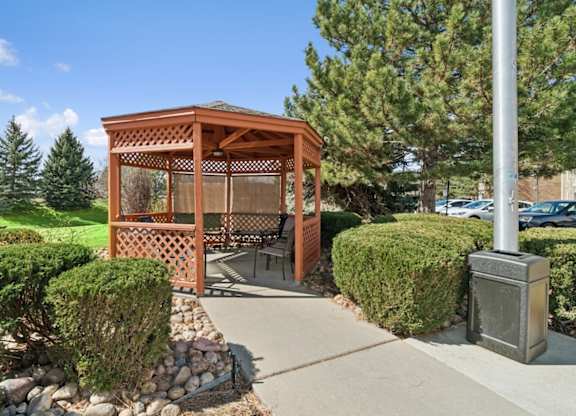 A wooden gazebo is surrounded by green bushes and a stone wall.