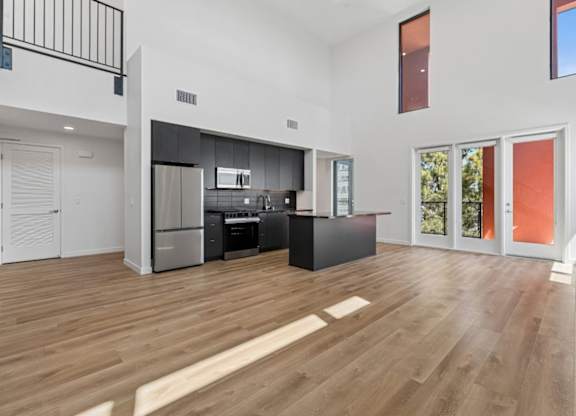 A kitchen with a refrigerator, oven, and sink is in the middle of a spacious room with wooden flooring.