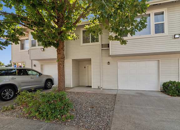 A grey SUV is parked in front of a two-story beige house with a tree in front.