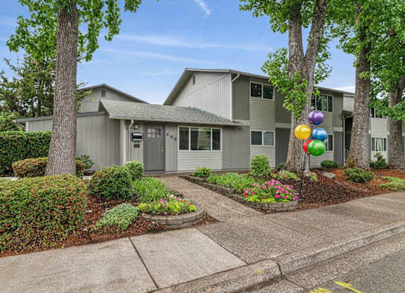 A house with a grey facade is surrounded by greenery and has a bunch of balloons tied to a tree.