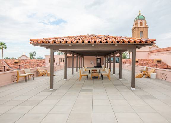 A patio with a roof and chairs is in front of a building with a tower.