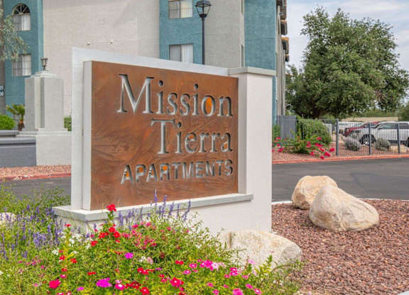 A sign for Mission Tierra Apartments is surrounded by flowers and rocks.