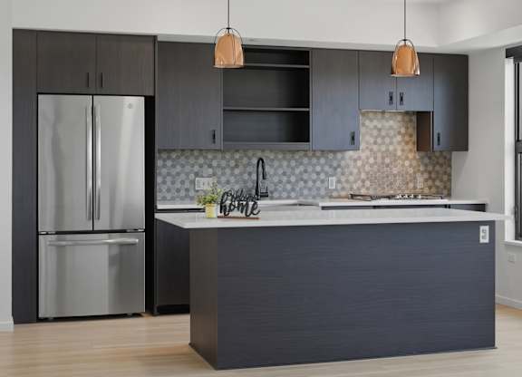 A modern kitchen with a dark grey island and a stainless steel refrigerator.