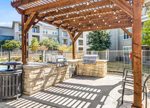 A wooden pergola is over a stone wall with a bench and a fire pit.