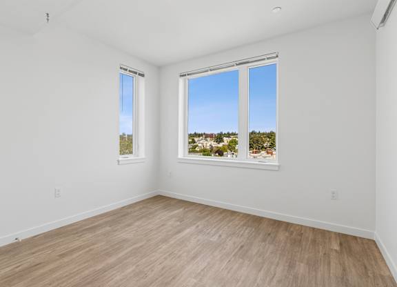 A room with white walls and wooden flooring, featuring two windows with a view of a residential area.