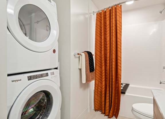 A white Whirlpool washing machine sits next to a bathroom with orange curtains.