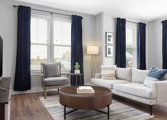A living room with a grey sofa, a wooden coffee table, a grey armchair, a TV, and a window with blue curtains.