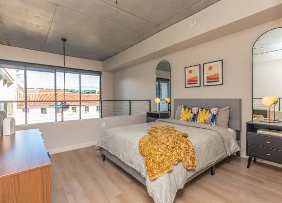 A bedroom with a bed, nightstand, and a window with a view of a red roofed building.