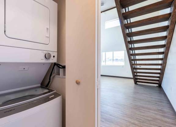 A white dishwasher is in a narrow kitchen next to a wooden staircase.