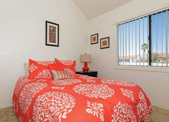 A bed with a red and white comforter is in the foreground of a room with a window.